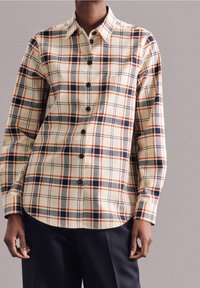 Camicia plaid beige con accenti neri e arancioni, maniche lunghe, chiusura frontale con bottoni e orlo curvo, realizzata in un tessuto texturizzato.