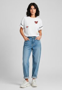 Witte t-shirt met korte mouwen en kleurrijke grafische prints, gecombineerd met lichtblauwe jeans met opgerolde mouwen en witte sneakers. Eenvoudig, casual ontwerp.
