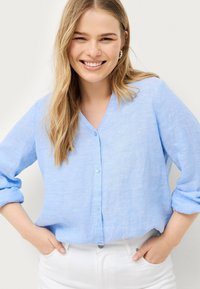 Chemise boutonnée en lin bleu clair avec manches longues et col en V. Le tissu a une finition texturée et un aspect légèrement froissé.