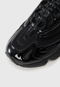 Black athletic shoe featuring a combination of mesh and shiny patent materials, rounded toe, and textured accents along the sides.