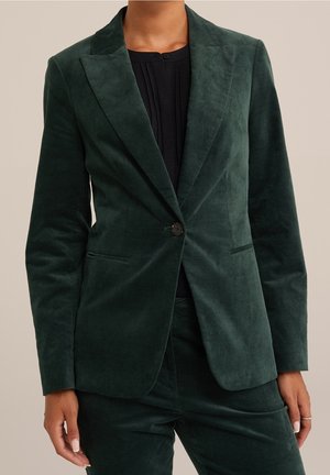 Skræddersyet mørkegrøn fløjlsblazer med enkelt knap, båret over sort top, kombineret med matchende fløjlsbukser, hænder afslappet langs siderne.