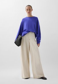Oversized paarse sweatshirt met lange mouwen gecombineerd met beige wijde broekspijpen. Model houdt een zwarte tas vast. Soepele stof, relaxte pasvorm.
