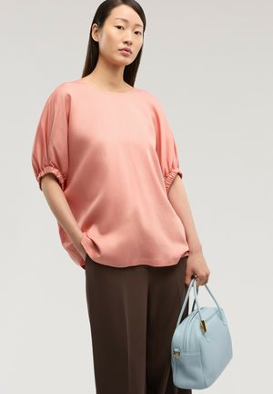 Frau trägt eine lockere rosa Bluse mit gerafften Ärmeln, dunkelbraune Hose und hält eine hellblaue Handtasche vor einem schlichten Hintergrund.