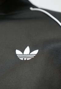 Juoda sportinė apranga su balta Adidas logotipu. Logotipą sudaro trys lapeliai ir horizontalios juostelės, esančios ant tekstūruoto audinio fono.