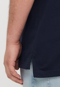Marineblauwe poloshirt van een textuurstof, met een zijvent aan de zoom. Hand in lichtblauwe jeans onderaan weergegeven.