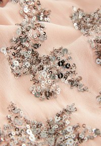 Tissu beige orné de grappes de sequins argentés brillants cousus en motifs floraux, créant une surface texturée et réfléchissante.