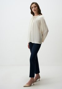 Blouse en crème fluide avec un col froncé et des manches longues, associée à un pantalon bleu marine ajusté et des escarpins beiges à bout pointu.
