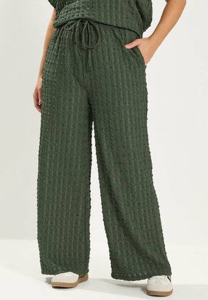 Femme portant un pantalon large texturé vert foncé à taille coulissante, une main dans la poche, associé à des baskets blanches.