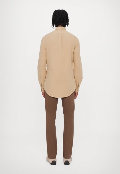 Camisa de manga longa beige com hem círculo e botões nos punhos, combinada com calças ajustadas castanhas e calçado de cor clara.