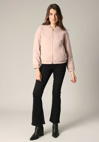 Lichtroze bomberjack met een rits aan de voorkant, geribbelde manchetten en zijzakken, gecombineerd met uitlopende zwarte jeans en enkellaarzen.