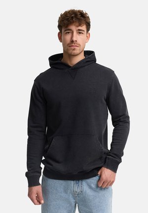 Ung mand med krøllet hår iført en sort hættetrøje og lyseblå jeans, stående mod en ensfarvet hvid baggrund.