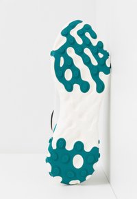 Semelle d'une basket blanche avec des motifs en caoutchouc bleu sarcelle et des plots de traction circulaires sur une surface texturée.