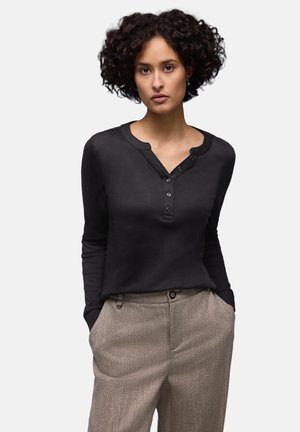 Kobieta z kręconymi włosami, ubrana w czarną, długorękawową, zapinaną na guziki koszulę oraz beżowe spodnie, stojąca z rękami w kieszeniach na jednolitym tle.