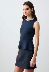 Top peplum blu navy con scollatura decorata da pietre, abbinato a una gonna blu navy a righe sottili. Tessuto liscio con silhouette strutturata.