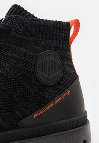 Chaussure montante tricotée noire avec un matériau texturé, un onglet de traction orange et un logo rond noir. Comprend une semelle en caoutchouc avec une finition à motifs.
