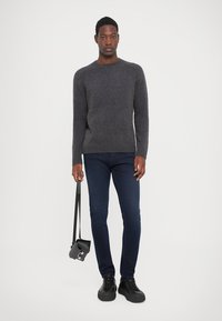 Man staand, gekleed in een grijze trui, donkerblauwe spijkerbroek, zwarte sneakers, en houdt een zwarte crossbody tas vast aan de schouderriem.