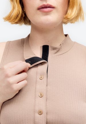 Person mit kurzen blonden Haaren, die einen beigen gerippten Top mit Knöpfen und einem schwarzen Klettverschluss in der Nähe des Halses schließt.