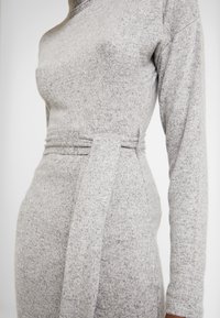 Gros plan d'une personne portant une robe en maille grise cintrée à manches longues avec une ceinture en tissu assorti nouée à la taille.