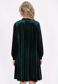 Rochie din catifea de culoare verde închis, cu mâneci lungi, fustă evazată și decolteu rotunjit, având o textură moale și netedă.