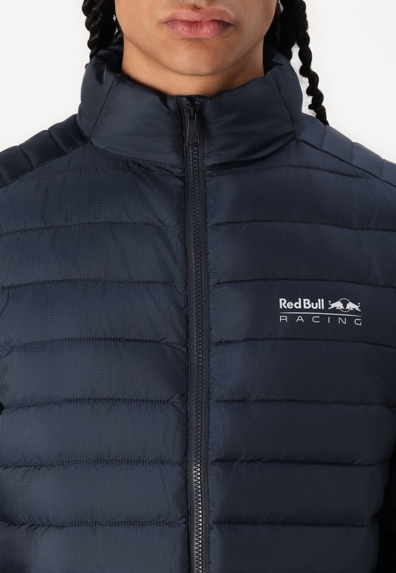 Marine pufferjack met een hoge kraag, verticale stiksels, volledige rits en het Red Bull Racing-logo op de linkerborst. Gladde, lichte stof.