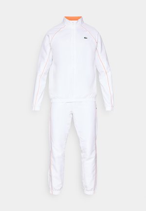 Witte sportieve trainingsoutfit met oranje accenten, bestaande uit een jack met rits en hoge kraag en elastische manchetten, en bijpassende tapering broek.