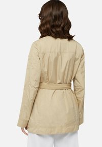 Giacca beige con cintura in vita, caratterizzata da una texture liscia e un taglio dritto. Il retro presenta un design semplice e sartoriale.