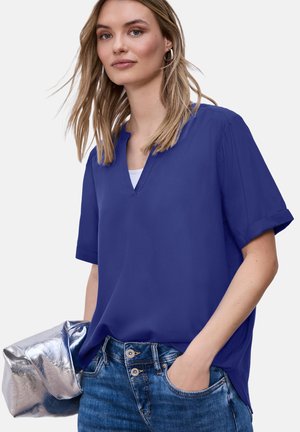 Femme portant un chemisier bleu à manches courtes et col en V, ainsi qu'un jean, tenant une pochette métallique argentée, une main dans la poche.