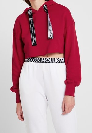 Sudadera corta roja con cordones de marca en negro y blanco, combinada con pantalones de chándal blancos que tienen una cinturilla a cuadros en negro.
