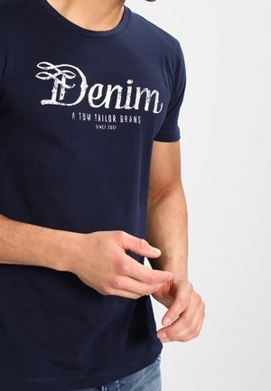 Mann trägt dunkelblaue T-Shirt mit dem Text „Denim Tom Tailor Brand Since 2007“, richtet die Finger, dargestellt vom Hals bis zur Taille vor einfarbigem Hintergrund.