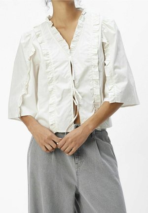 Kvinde iført en hvid bluse med flæser, trekvartlange ærmer og bindelukninger, sammen med løse grå bukser.