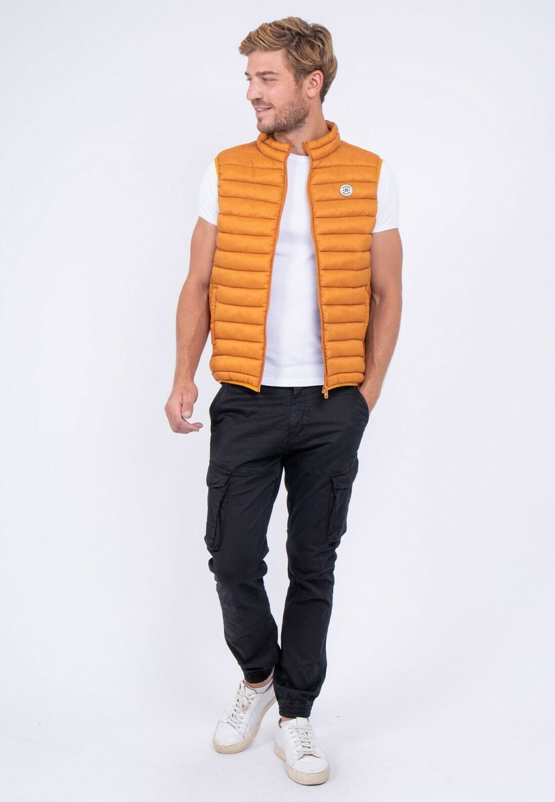 Gilet rembourré orange avec des coutures horizontales, col montant et fermeture éclair à l'avant, porté sur une chemise blanche et associé à un pantalon cargo noir et des baskets blanches.