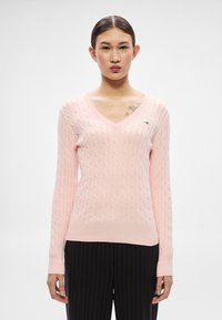 Rozā, kabelu raksta džemperis ar V veida kaklu, garām piedurknēm, ribota apakša un nelielu logo kreisajā krūtīs.