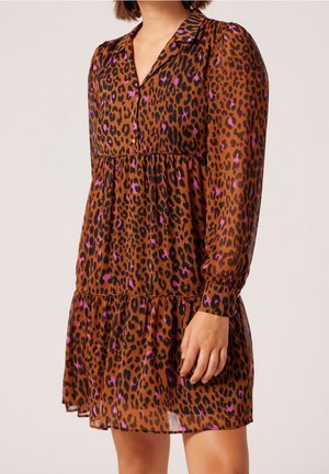 Kobieta ubrana w brązową sukienkę w panterkę z fioletowymi akcentami, z długimi rękawami, kołnierzykiem i warstwową spódnicą, stojąca na jednolitym tle.