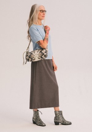 Mujer con cabello largo y gris y gafas, que lleva un top azul, falda marrón, botas con estampado de serpiente y lleva un bolso de hombro a juego con estampado de serpiente.