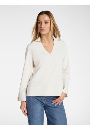 Vrouw die een wit V-hals trui met lange mouwen en een blauwe spijkerbroek draagt, poseert met één hand op haar heup en kijkt opzij.