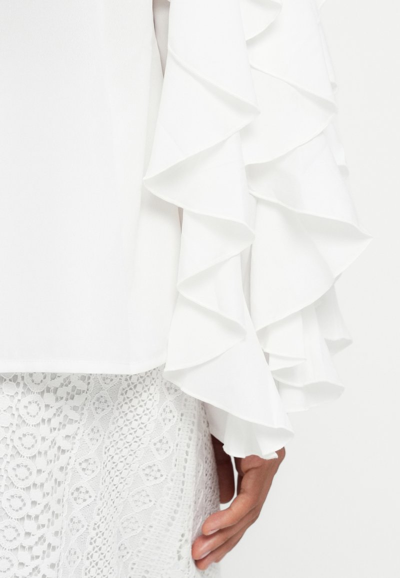 Weißes, gerafftes Blusensleeve mit geschichteter, geschwungener Stoffstruktur, kombiniert mit einem weißen Spitzenboden, der ein geometrisches Muster aufweist.