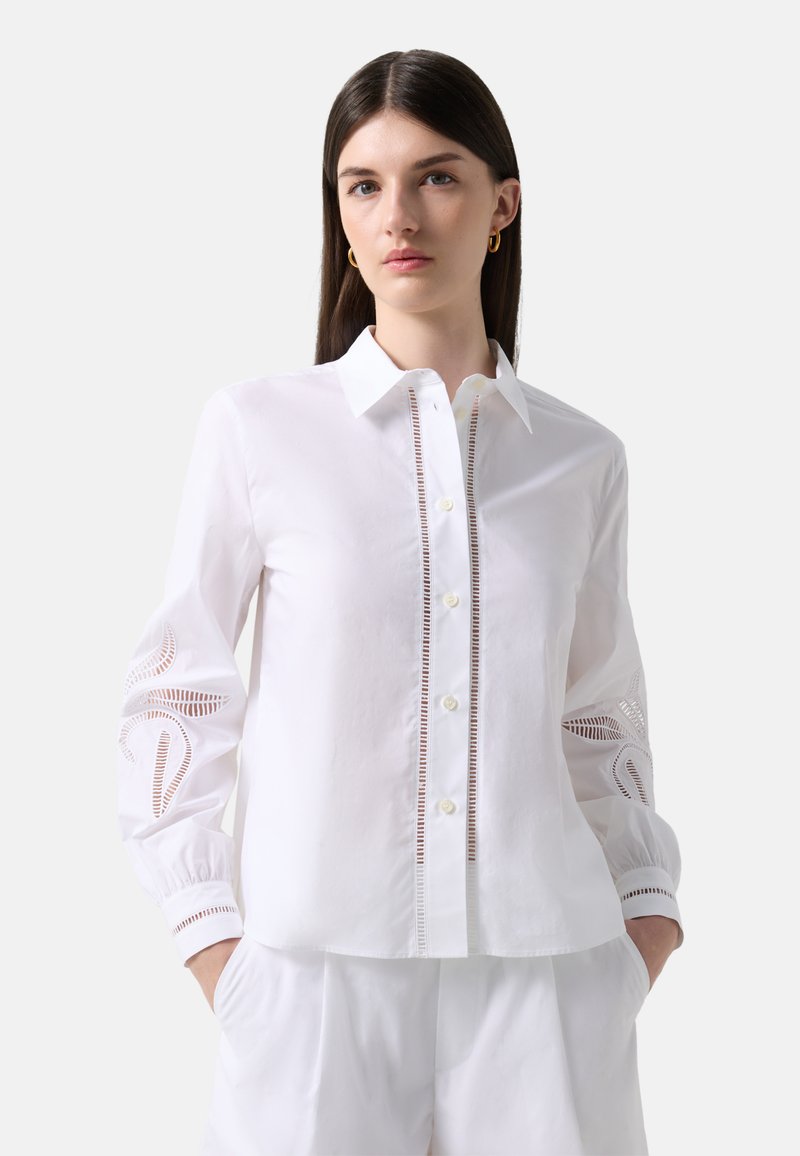 Kobieta ubrana w białą koszulę na guziki z koronkowymi wycięciami na rękawach i z przodu, ubrana w białe spodnie, stojąca na jednolitym tle.