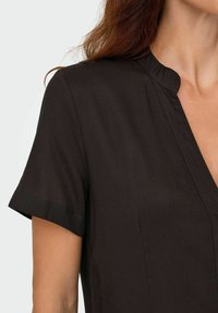 Femme portant un blouse noire à manches courtes avec un col à petit encoignure, cheveux bruns visibles, recadrée des épaules jusqu'au milieu de la poitrine sur fond uni.