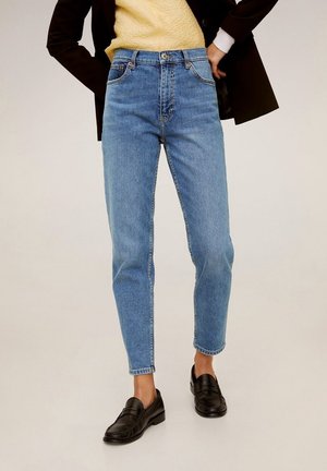 Persoon die lichtblauwe jeans met hoge taille draagt, zwarte loafers, een gele getextureerde top en een zwarte jas met handen in de zij.
