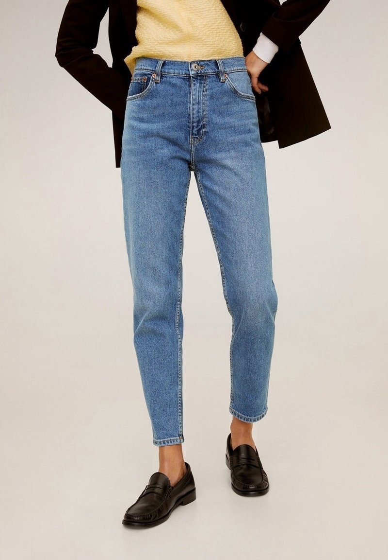 Personne portant un jean taille haute bleu clair, des mocassins noirs, un haut jaune texturé et une veste noire, les mains sur les hanches.