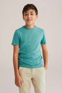 Tirkīza krāsas kokvilnas T-krekls ar īsām piedurknēm un apaļu kaklu, apvienots ar gaiši beige biksēm. Gluda tekstūra, ikdienišķa piegriezne, minimāls dizains.