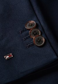 Τρία σκούρα καφέ κουμπιά με την επιγραφή "Tommy Hilfiger" στη μανσέτα ενός ναυτικού μπλε κουστουμιού με μικρό ορθογώνιο καρφί με κόκκινο, λευκό και μπλε λογότυπο.