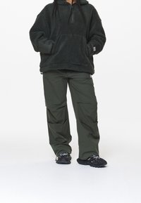 Ciemnozielony, futrzasty sweter z suwakiem o ćwierć długości i dużą przednią kieszenią, w zestawieniu z luźnymi, ciemnozielonymi spodniami cargo. Czarne buty sportowe.