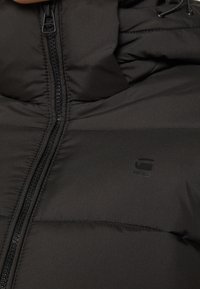 Veste rembourrée noire avec texture matelassée, dotée d'une fermeture éclair à l'avant et d'une capuche réglable avec cordon. Logo discret imprimé sur la poitrine.