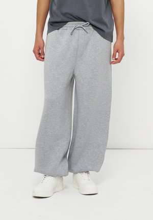 Osoba nosząca luźne, jasnoszare spodnie dresowe z sznurkiem w talii oraz białe trampki, stojąca na tle jednolitego białego tła.