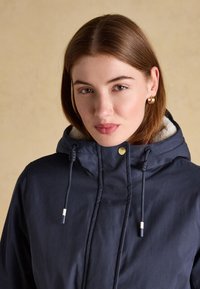 Giovane donna con capelli castani indossa orecchini a cerchio dorati e una giacca con cappuccio blu navy con fodera in pile e cordini.