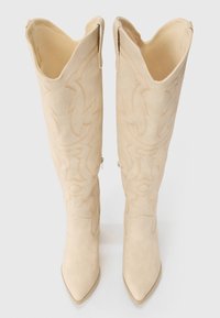 Bottes crème montantes avec des orteils pointus, des coutures décoratives et un design western ; elles présentent une texture en daim doux et des fermetures éclair sur les côtés.