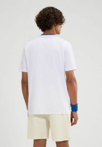 Weißes T-Shirt mit kurzen Ärmeln und blauen Akzenten am Kragen, kombiniert mit hellgelben Shorts. Der Stoff wirkt glatt und hat minimale Designdetails.