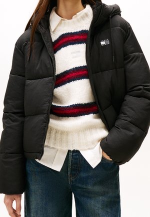 Vrouw draagt een zwarte donsjas over een witte gebreide trui met rode en marineblauwe strepen, een wit overhemd met kraag en een blauwe spijkerbroek.