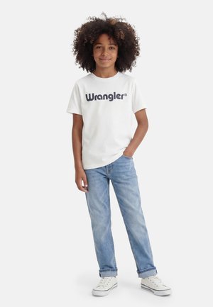 Laps lokkis juustega, kellel on seljas valge Wrangler T-särk, helesinised teksad ja valged spordijalatsid, seisab ühe käega taskus.